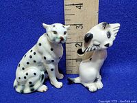 Two porcelain cat figurines placed next to a ruler showing size around 3.5-4 inches, white with black spots and markings, sitting poses, no visible damage.