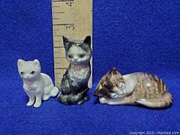 Three different porcelain cat figurines standing and lying in front of ruler for size reference. White cat about 2 inches tall, medium grey/black cat about 4 inches tall, brown tabby cat lying down around 3 inches long.