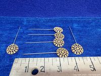 Six vintage gold tone filigree hat/scarf pins arranged horizontally with ruler showing measurement scale to indicate length of about 3-4 inches.