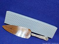 Pie or cake server with polished triangular blade and ornate floral patterned handle placed before its light blue patterned box on a blue background.