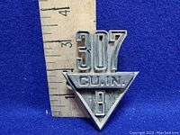 Front of the chrome car emblem showing '307 CU.IN. V8' with a ruler indicating size, approximately 2.5 inches tall.