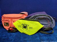 Three bags displayed on a dark blue background: bright yellow smaller fanny pack with Garfield Survival Kit logo, orange large fanny pack, purple and gray crossbody bag.