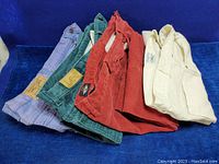 Five pairs of vintage pants laid side by side including purple Jordache, teal Lee jeans, red corduroy pants, red Dockers corduroy, cream Chic pants on blue background.