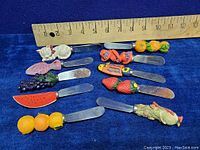 Photo showing 12 vintage appetizer or butter spreaders with decorative painted handles including animals, fruits, and characters. A wooden ruler above showing measurements around 3 to 4.5 inches long.
