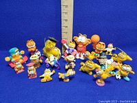 Photo shows 18 vintage Garfield vinyl figures, various costumes and activities, arranged in group against blue cloth background with measuring ruler behind.