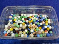 Top-down view showing a large number of assorted vintage marbles in a plastic container. Various colors, patterns, and sizes visible.