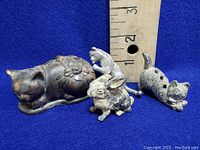 Front view of four metal animal figurines against blue background and ruler for size: three cats and one rabbit.