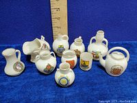 Ten miniature Goss crestware porcelain items arranged with a ruler for size reference showing various heraldic crests, all in excellent condition.
