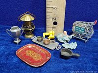 Miniature vintage items next to ruler for size reference. Includes a small silver trophy, brass lantern, yellow sewing machine, baby doll with blue crochet outfit, crocheted bonnet, red and gold tray, small metal baking mold, metal tray, frying pan, decorative brass pot, and shopping cart.