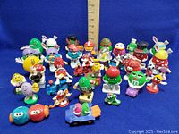 Thirty-four assorted vintage M&M’s collectible vinyl figures arranged in rows against a blue background with a ruler for scale