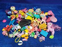 Pile of assorted small toys and toy accessories including small doll figure, My Little Pony figurines, and various colorful small toys spread on a blue surface.