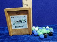 Photo of the wood box with sliding lid showing the label 'NPW 21 Marbles' surrounded by 21 marbles in assorted colors against a blue background with a ruler for size reference.