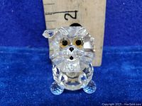 Close-up front view of clear crystal lion figurine showing faceted face, black nose, gold-tone eyes, and ears. Background ruler for size reference.