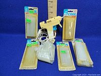 White hot glue gun with power cord surrounded by several unopened packages of hot glue sticks on a blue background with a ruler for scale.