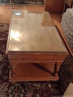 Side angle view of the rectangular solid oak coffee table with a glass top showing wood grain and turned legs, drawer beneath the top, and a bottom shelf.