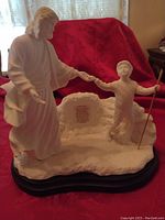Front view of white porcelain figurine showing robed man reaching to young boy with walking stick on a rock detailed base, mounted on wooden base.