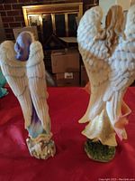 Back view of two Lenox angel figurines showing detailed textured wings and painted bases on red cloth background.