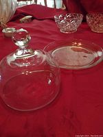 Glass butter dish with clear dome lid showing knob handle, placed on red fabric with other glassware partially visible.