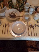 Photo of place setting with dinnerware and silverware for one place setting, showing forks, spoons, knives, plates, bowls, cups.