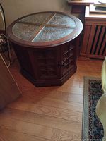 Octagonal wooden drum table with fold-out door and decorative inlaid glass top with floral patterns, placed on wood floor next to furniture.