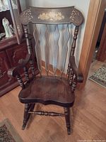 Front view of dark stained solid wood rocking chair with carved floral decoration on the top backrest panel, contoured wooden seat, turned armrests and legs.