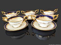 Five porcelain double-handled bouillon cups with matching saucers set on black surface showing cobalt blue and gold gilt decoration.