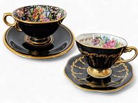 Two fine china tea cups and saucers with black floral designs and gold trim shown from the front
