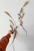Photo showing two copper and brass wheat stalk wall art pieces held against a wall for scale, highlighting size and detail.