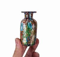 Photo of a small 3-inch Murano glass bud vase held between fingers showing detailed multicolored millefiori cane patterns with gold foil inclusions on the surface.