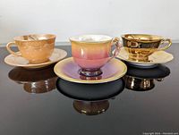 Three vintage tea cup and saucer sets displayed on a reflective black surface, showing colors ranging from peach lustre, ombre pink to gold, highlighting shapes and lustre finishes.