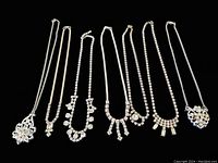 Top view of all 7 silver-tone rhinestone necklaces laid out on black background showing varying chain and pendant designs.