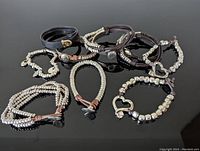 Top down view of 9 vintage bracelets arranged on black surface showing varied styles including leather, cords, and silver beads with button details.