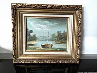 Front view of the oil painting depicting a boat on a waterway with scenic landscape, in an ornate wood frame.