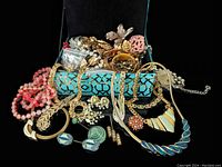 Jewelry box opening with costume jewelry overflowing, showing various gold-tone bracelets, coral pink bead necklaces, and assorted brooches and earrings