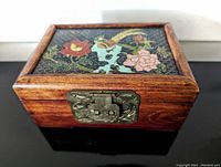 Front view of the closed rectangular box showing the rosewood frame, handpainted bird and flower design on black textured background, and brass embossed lock hardware.