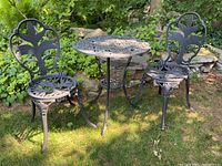 Two black aluminum chairs and a round metal table outdoors on grass, showing overall set condition and design.