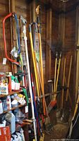 Photo of garden hand tools including two bow saws, rakes, shovels, brooms, a tiller, and other tools stored against wooden wall.