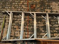Full length view of the aluminum orchard ladder laid horizontally against a brick wall with visible bracing supports and wide rungs.