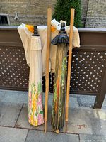 Pair of two tall folded paper umbrellas with bamboo slats leaning against a wooden fence with two cotton bags behind them.