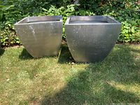 Pair of square galvanized metal planters placed on grass outdoors against leafy background, showing tapered bottom shape and wear marks.