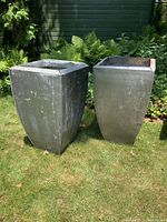 Pair of galvanized metal planters standing outdoors on grass with plants in background, showing overall shape and size