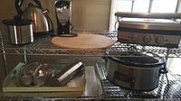 Wide view of kitchen appliances on metal shelving including kettle, can opener, pizza stone, Crock-Pot and trays.