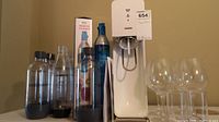 Photo showing SodaStream Source machine with five carbonation bottles and six glasses placed in front and beside.