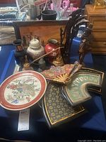 Overview of lot displaying the Satsuma peacock plate, Asian vases, fans, fan-shaped ceramic wall art, and carved wooden figurines on a blue tablecloth.