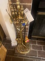 Full view of the brass fireplace tools set in stand showing poker, tongs, broom, shovel and attached match holder near a brick fireplace.