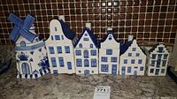 Seven pieces of hand-painted Delft blue ceramic canisters shaped as Dutch houses and a windmill, displayed side by side to show design and size difference.
