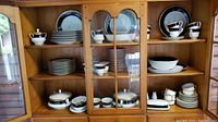 Wooden cabinet holding numerous plates, cups, and saucers arranged on shelves. Shows variety and quantity of china pieces for service of 12 plus additional items.