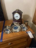 Set of three clocks displayed on wood surface: Pottery Barn triple face stainless steel clock (center with round clock, temperature, and humidity dials), Kensington wooden carriage style clock (arched top, brass feet) behind it, and small gold-toned Linden table clock on the right.