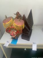 Two items on a white surface: a red Indonesian dragon mask with gold ornamental fringe and a dark carved wooden pagoda sculpture.