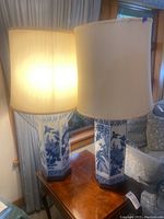 Two blue and white ceramic lamps with beige shades positioned on a wooden table by a window, showing overall appearance and style.